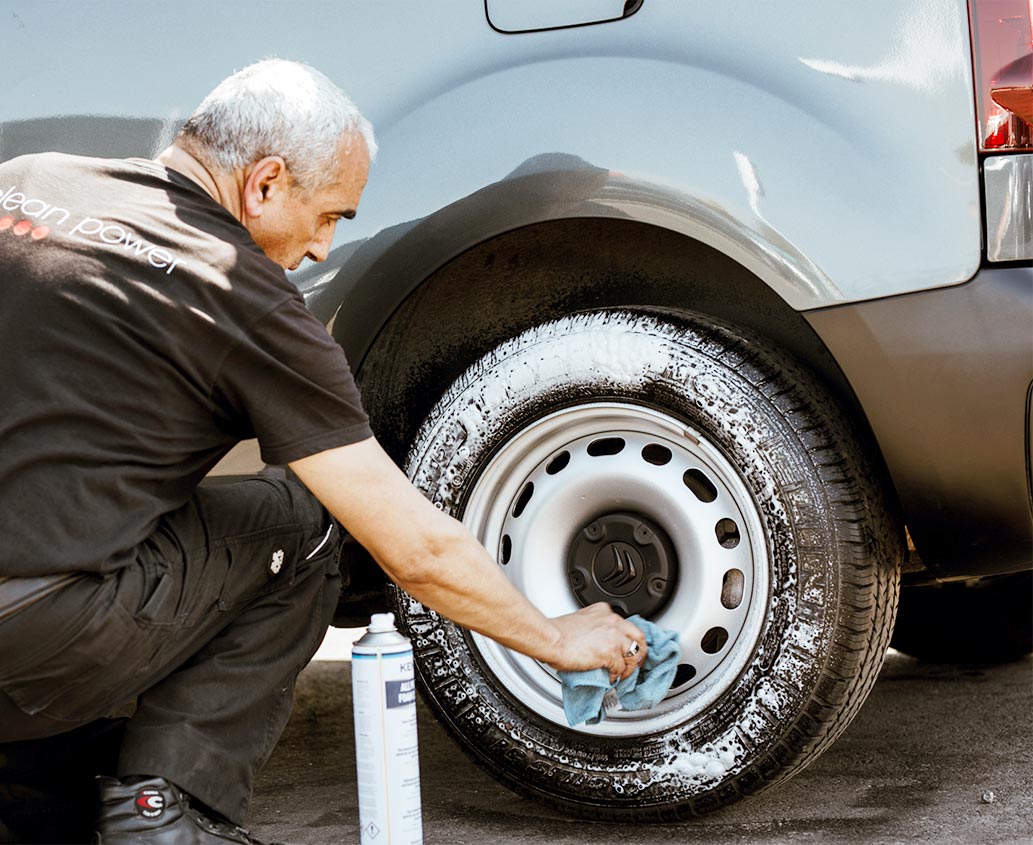Professionnels en train de nettoyer une jante de voiture avec du savon sur un parking.